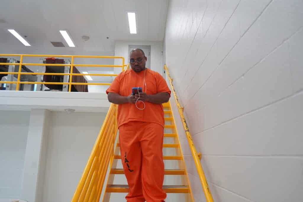 A man wearing orange prison attire stands on a stairway, looking down with a serious expression.